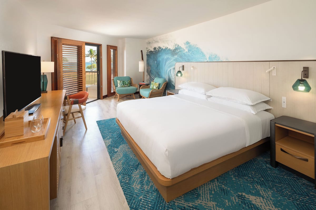 A hotel room with a bed, a desk, a TV, and a view of the ocean through the window.