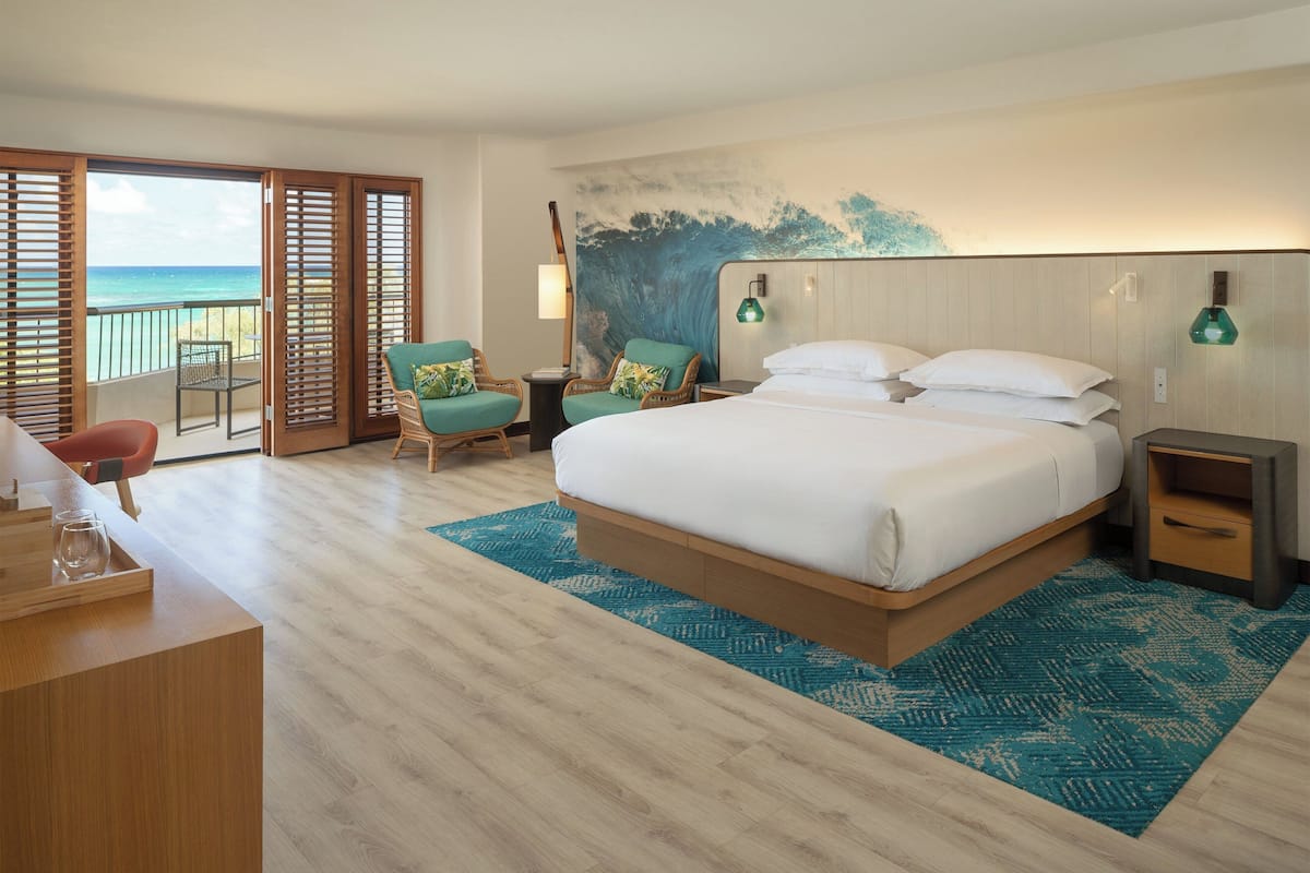 A hotel room with a large bed, a desk with a chair, a small table, and a view of the ocean through open sliding doors.