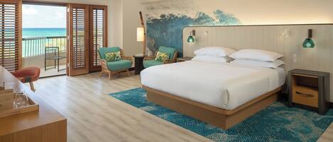 A hotel room with a large bed, a desk with a chair, a small table, and a view of the ocean through open sliding doors.