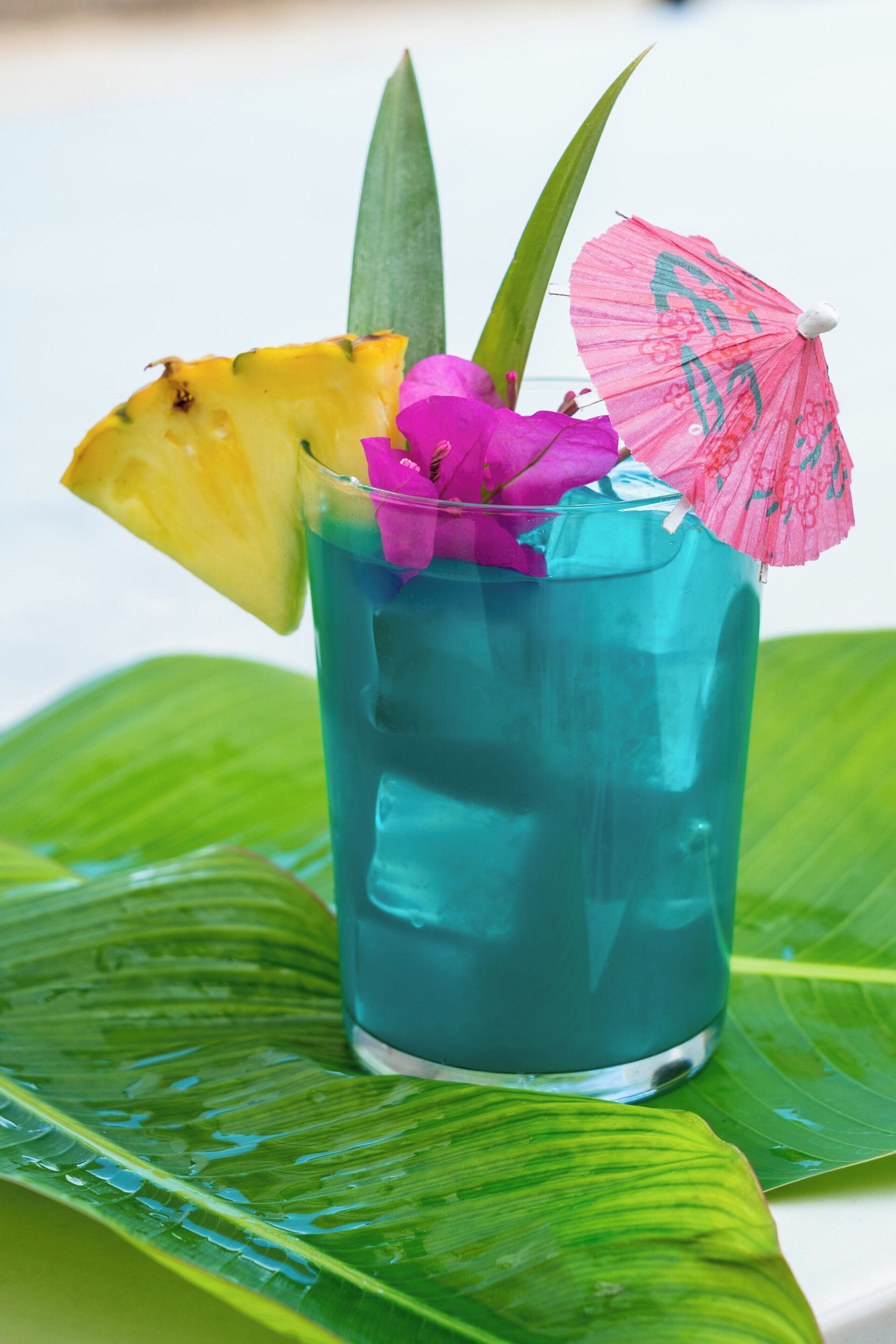 A clear glass with a blue liquid, garnished with a pineapple slice, pink flower, and a pink umbrella.