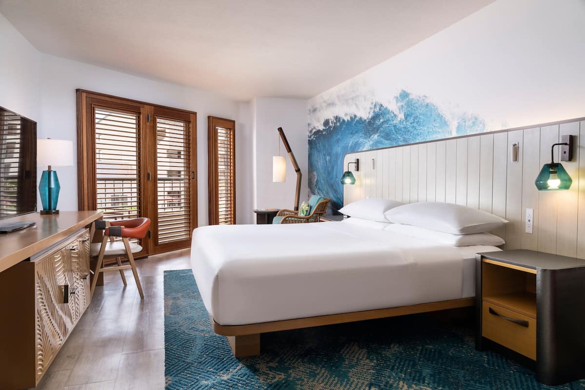 A hotel room with a bed, a desk, a chair, a nightstand, and a wall with a wave mural.