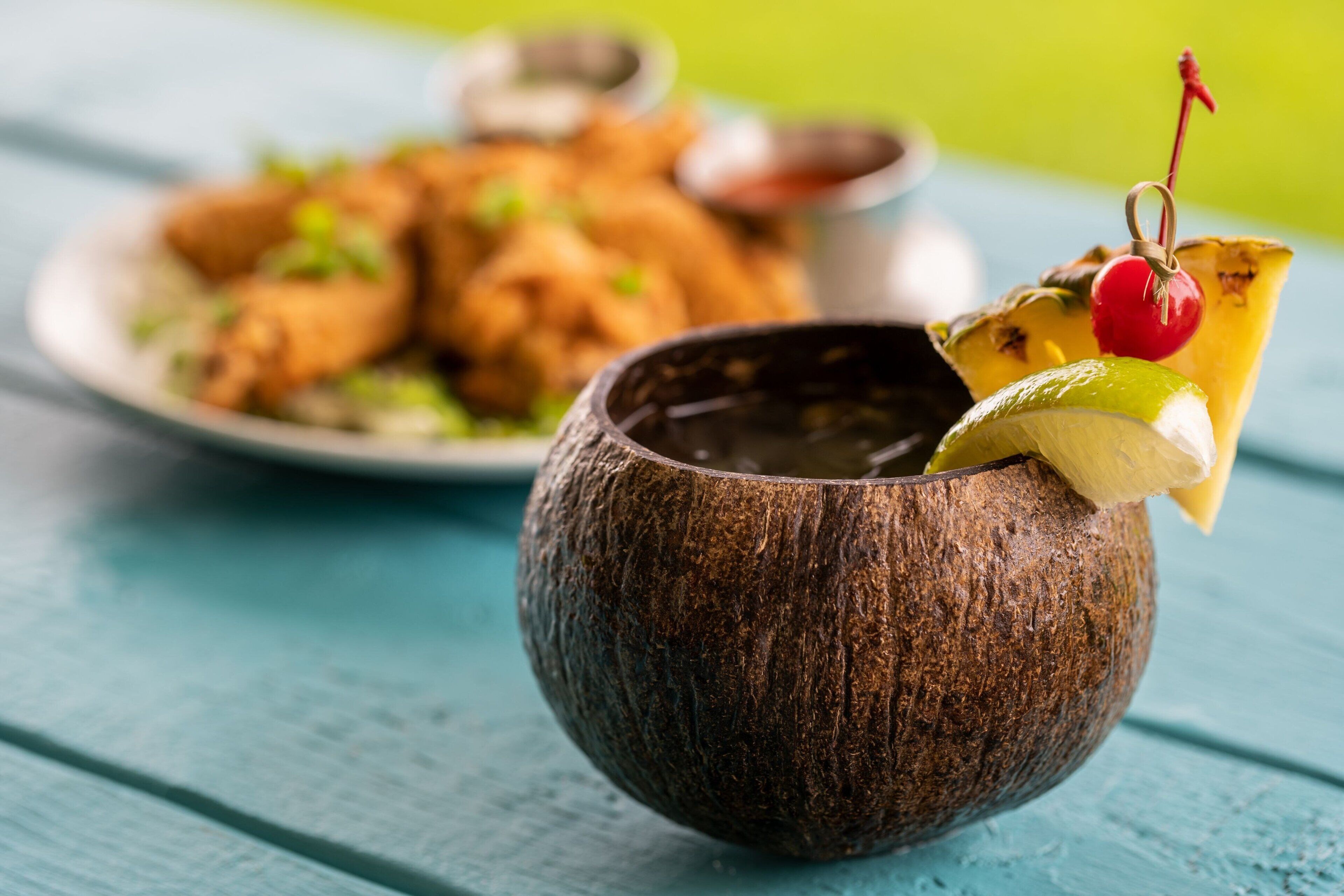 A wooden bowl with a lime wedge and a cherry on a cocktail glass, placed on a blue wooden surface.