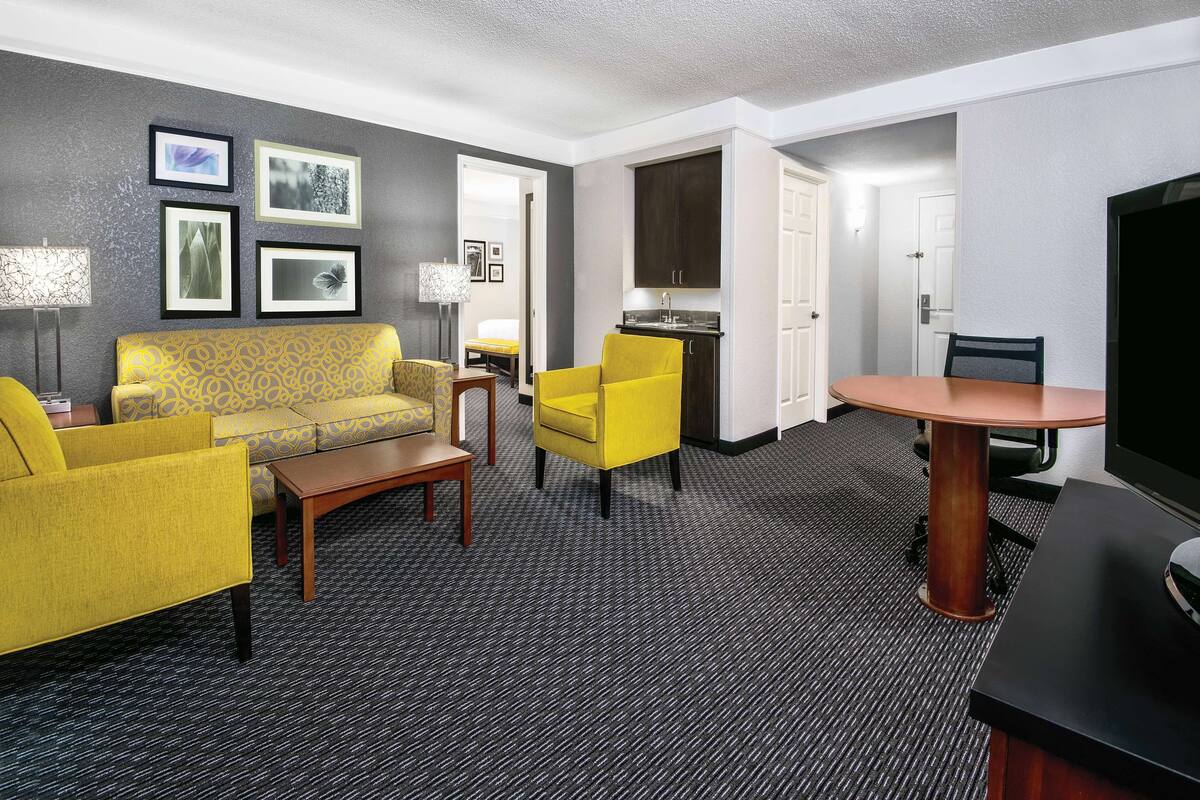 Une chambre d’hôtel comprenant un canapé, des fauteuils, une table à manger, un téléviseur, un coin cuisine et des tableaux encadrés au mur.