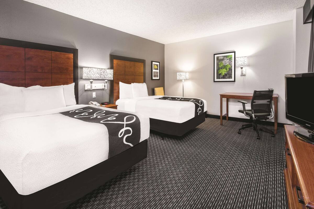 Une chambre d’hôtel avec deux lits, un bureau, une chaise, un téléviseur et deux lampes fixées au mur.