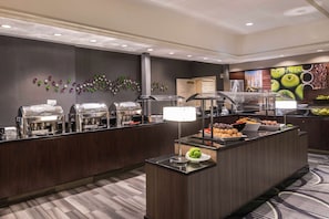 Ein Buffet mit verschiedenen Speisenstationen, einer Kaffeemaschine und einer Wand mit Frucht- und Kaffeebohnen-Mustern.