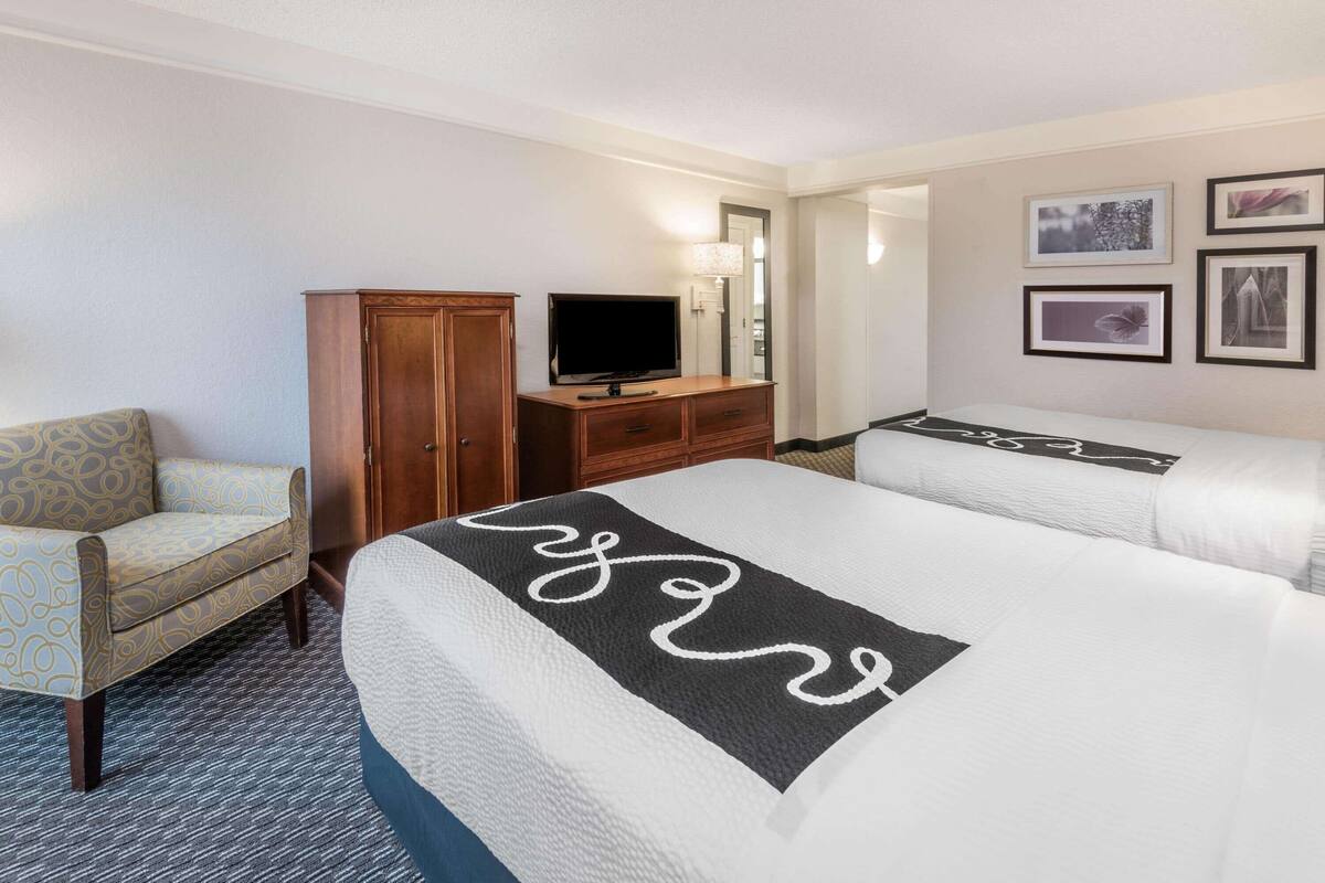 Une chambre d’hôtel avec deux lits, une télévision, une armoire, une chaise et des tableaux encadrés au mur.