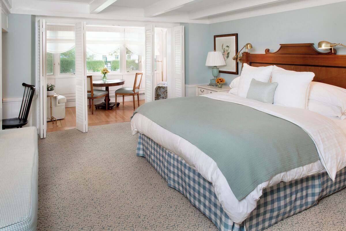 A bedroom with a large bed, a wooden headboard, a bedside table with a lamp, and a view through an open door to a dining area.