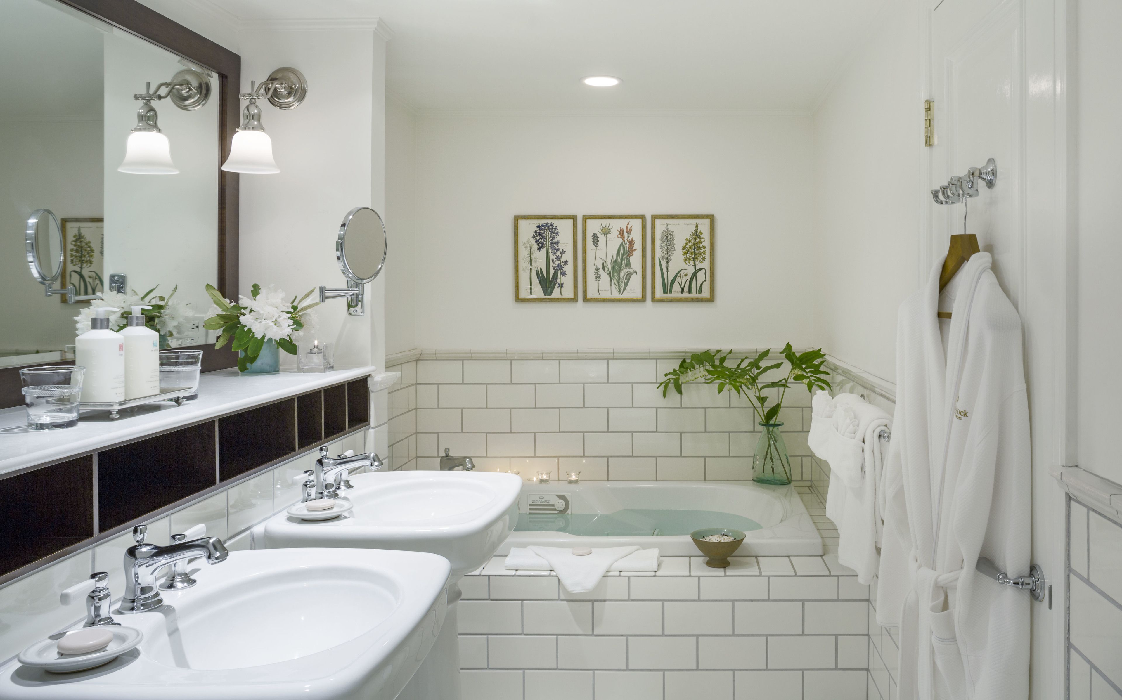 A bathroom with two sinks, a bathtub, and a large mirror. There are framed botanical prints on the wall and a potted plant.