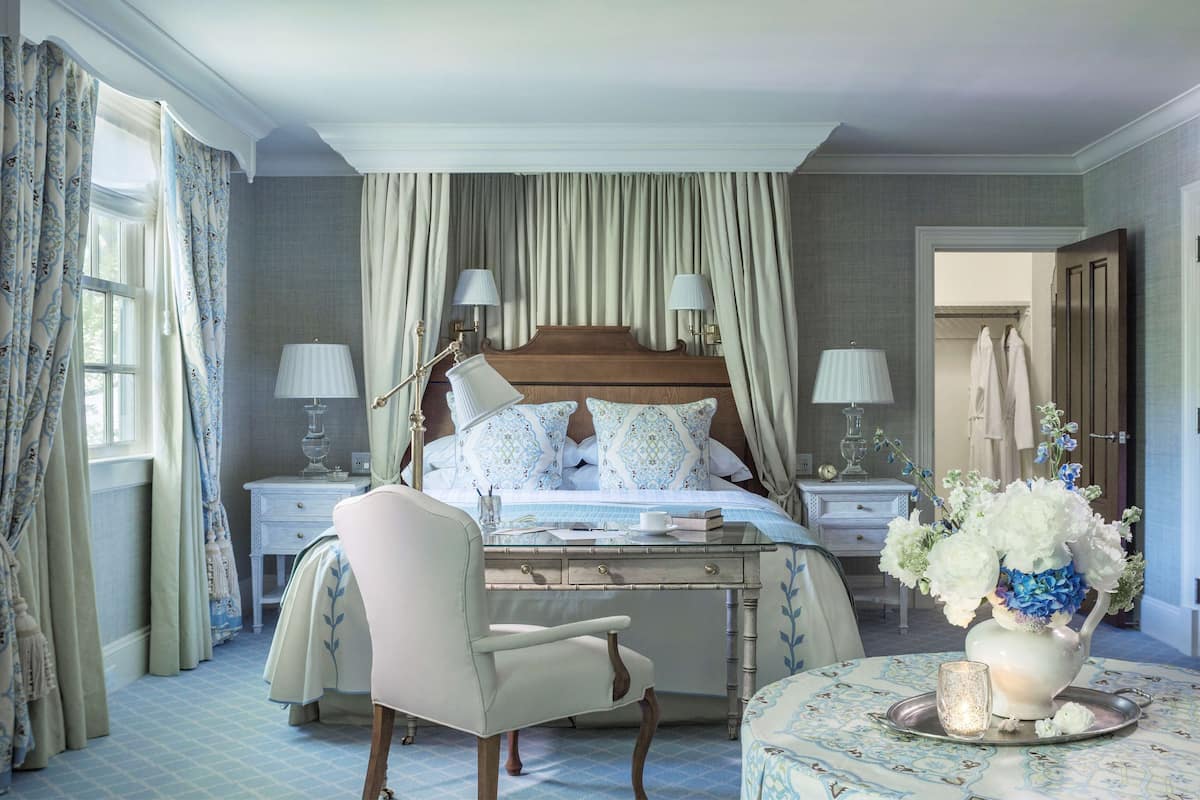 A bedroom with a large bed, two bedside tables with lamps, a chair, a bedside table with a vase of flowers, and a window with curtains.