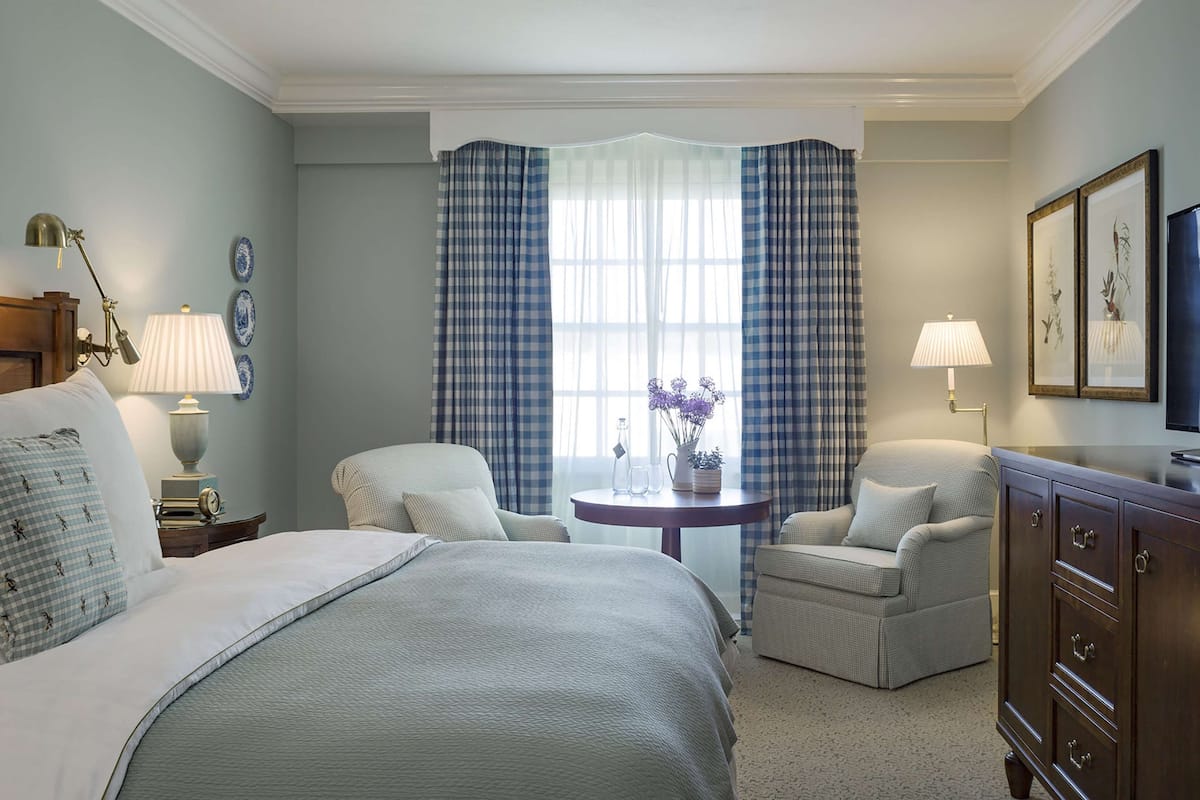 A hotel room with a bed, two armchairs, a small table, a TV, and a window with curtains.