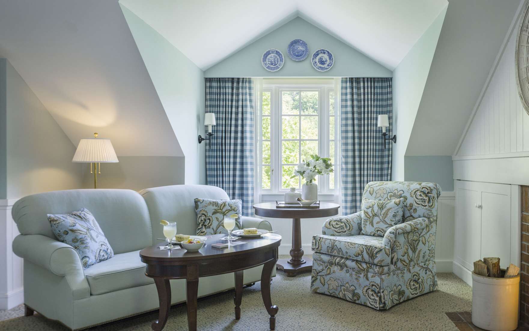 A cozy room with a light blue wall, a checkered window treatment, a floral armchair, a sofa, and a small round table with a vase and a lamp.