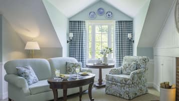 A cozy room with a light blue wall, a checkered window treatment, a floral armchair, a sofa, and a small round table with a vase and a lamp.