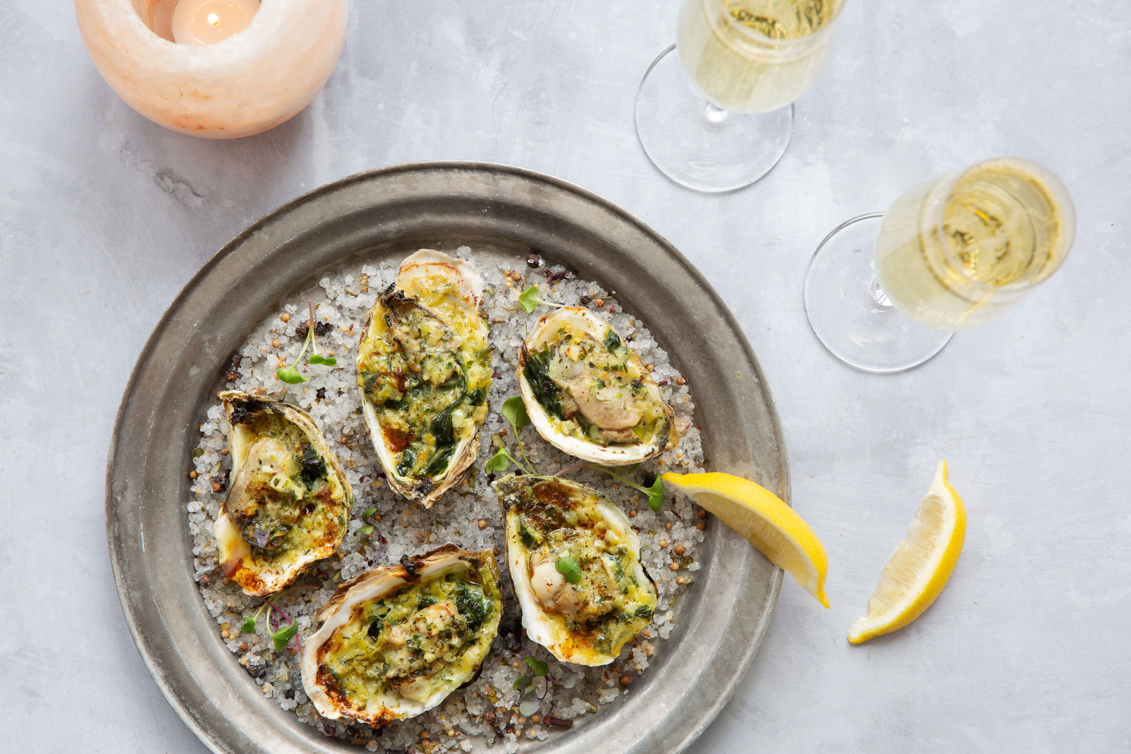 A plate of oysters on ice with a lemon wedge, accompanied by two glasses of white wine.