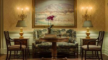 A room with a floral armchair, two side tables with lamps, a round table with a vase and books, and a large landscape painting on the wall.