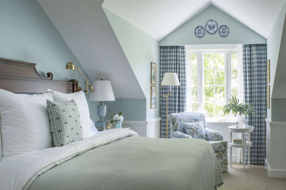 A bedroom with a bed, bedside lamps, a chair, a small table, and a window with curtains.