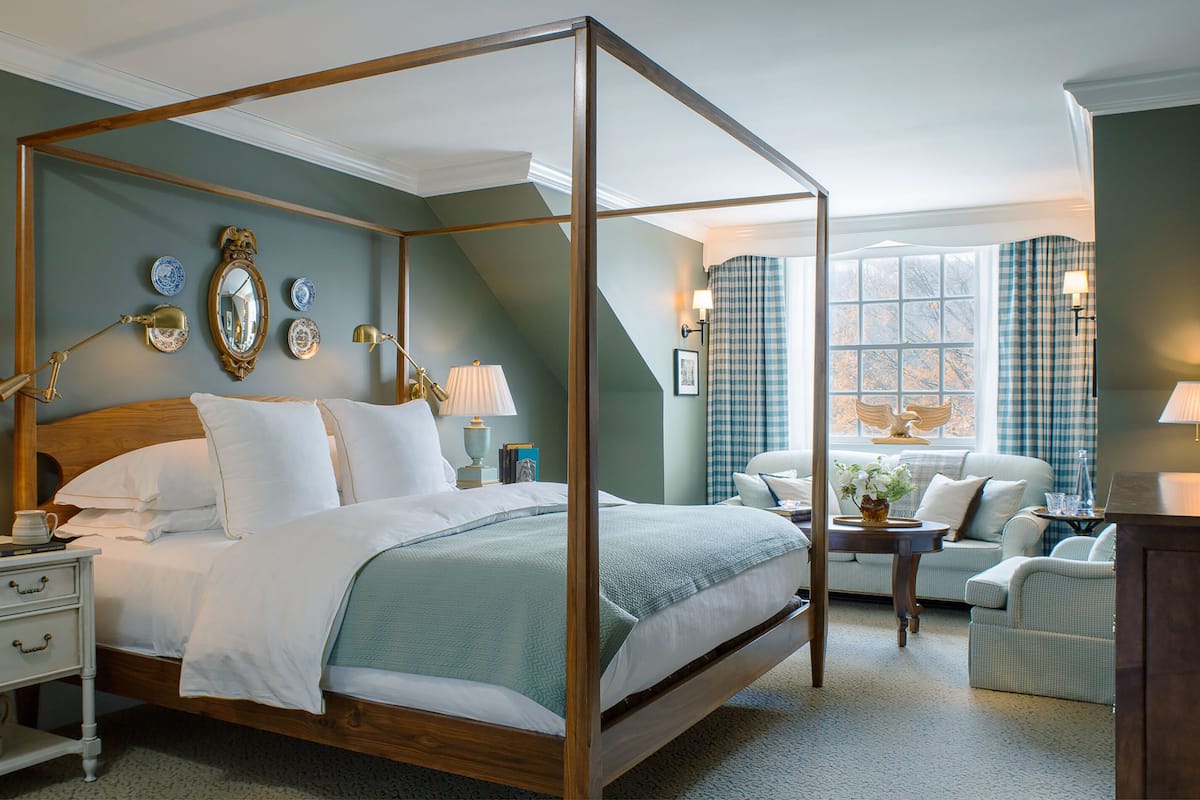 A four-poster bed with white linens, a nightstand with a lamp, and a seating area with a sofa and armchairs.