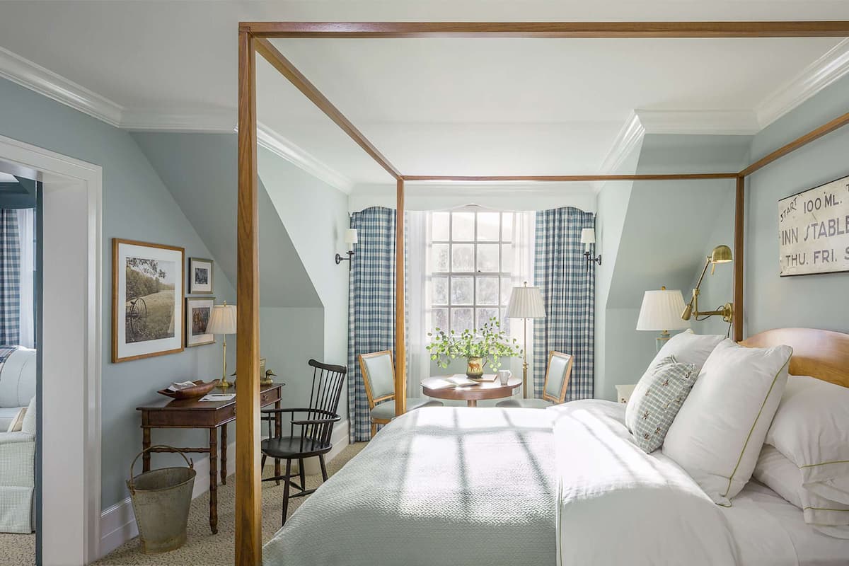 A bedroom with a four-poster bed, a desk with a chair, a window with curtains, and a wall-mounted lamp.