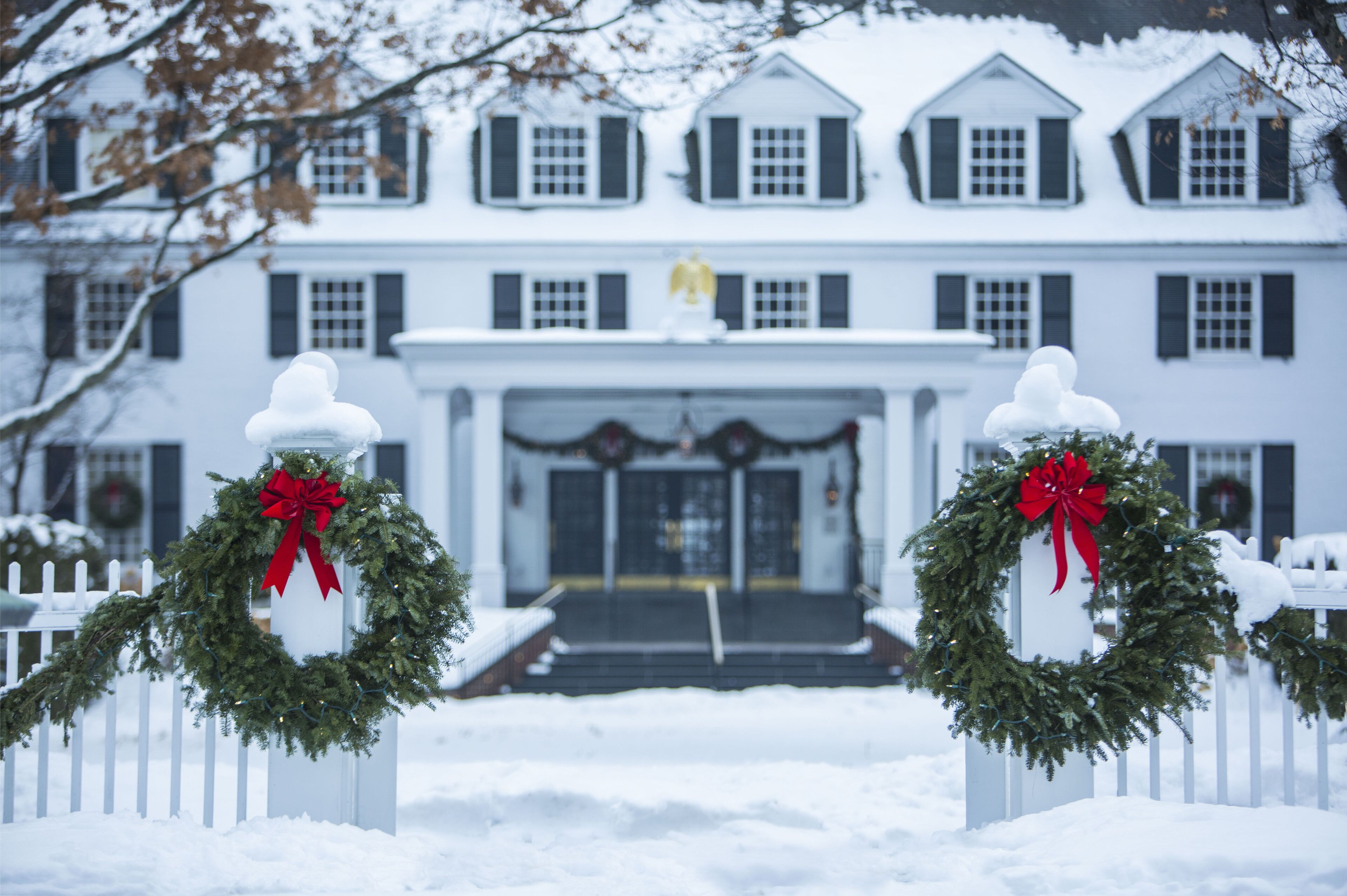 A large white house with a white picket fence, snow-covered steps, and two wreaths with red bows.