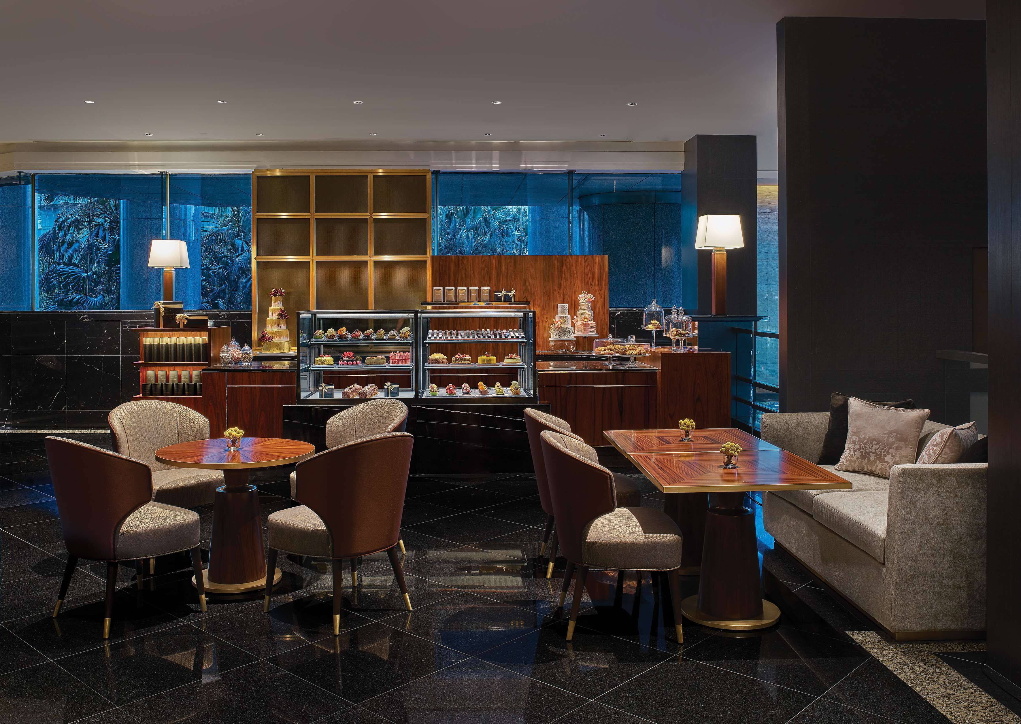 A modern hotel lobby with a central seating area, a dessert display, and a large window with a view.