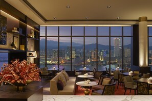 A modern hotel lobby with a large window offering a city view, a reception desk, and a seating area with tables and chairs.