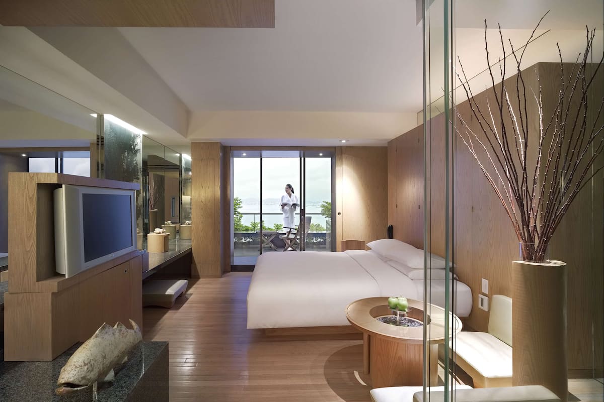 A modern hotel room with a large bed, a TV, a small seating area, and a view of the outdoors.