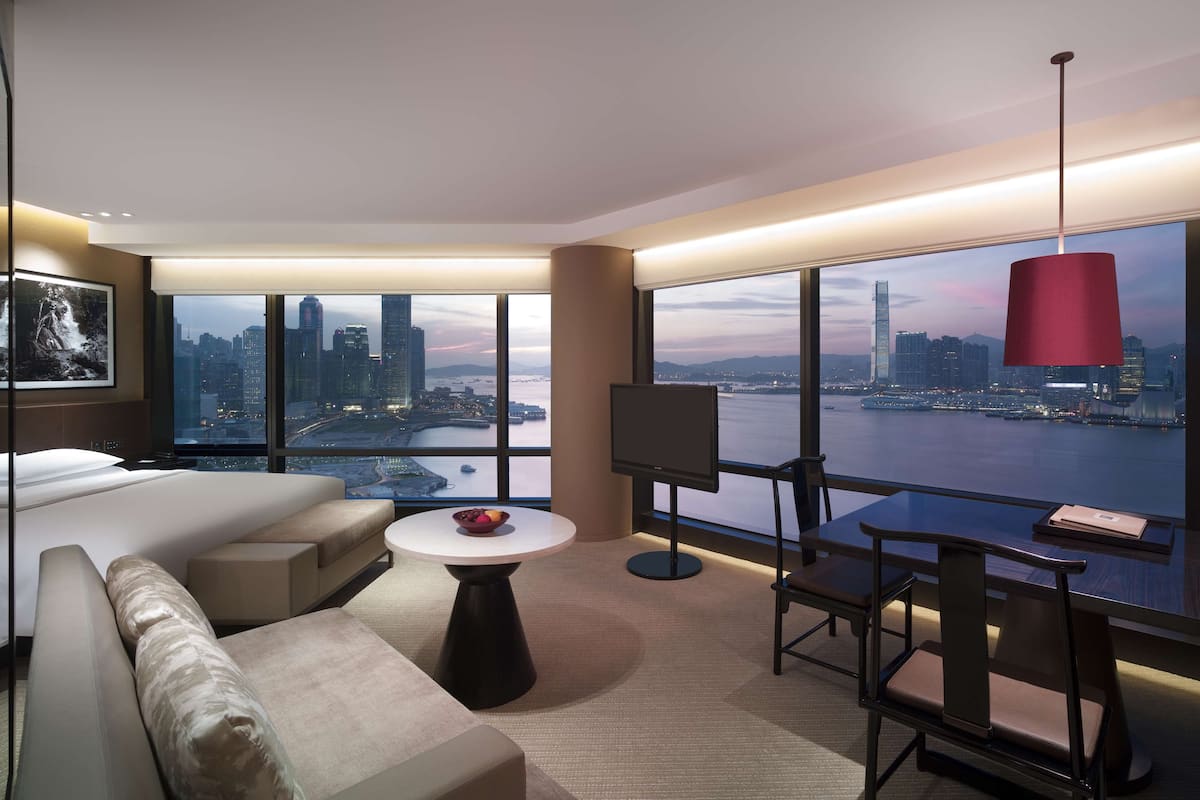 A modern hotel room with a large bed, a sofa, a dining table, and a TV. The room features a large window with a city view.