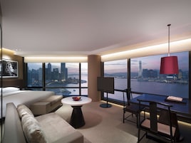 Quarto de hotel moderno com uma cama grande, sofá, mesa de jantar e televisão. O quarto tem uma janela ampla com vista para a cidade.