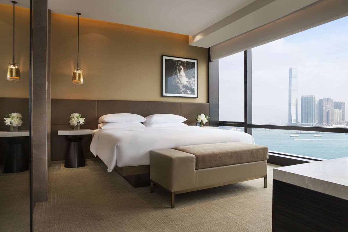 A modern hotel room with a large bed, bedside tables, a view of the city, and a cityscape through the window.