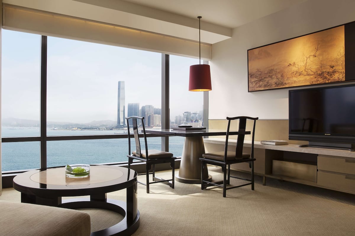 A modern hotel room with a large window offering a city view, a dining area with a round table, and a flat-screen TV.