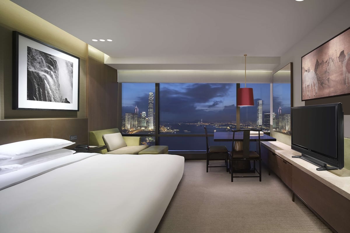 A modern hotel room with a large bed, a desk with a TV, a chair, and a view of a cityscape at night.
