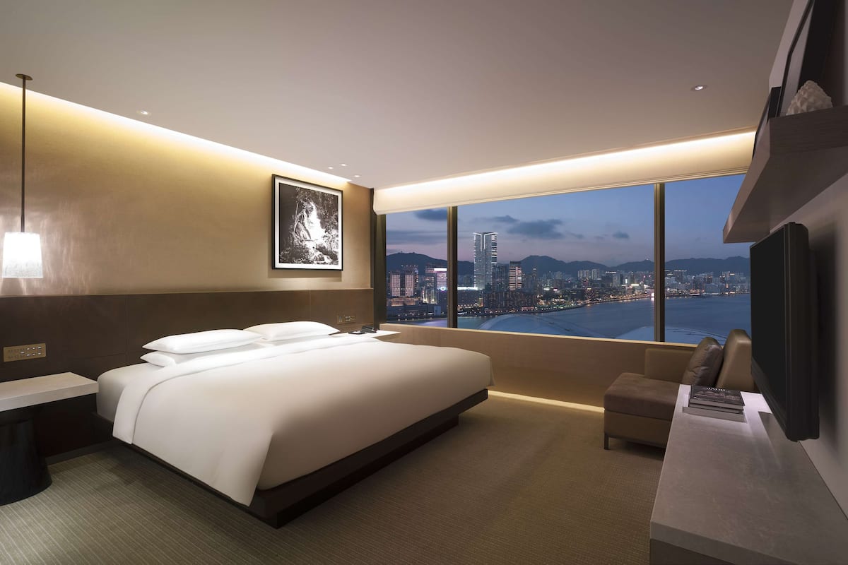 A modern hotel room with a large bed, a desk, a chair, and a city view through large windows.