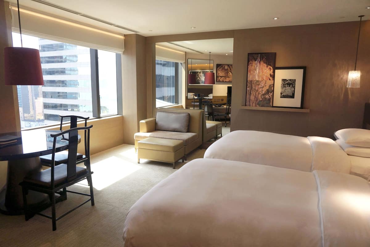 A hotel room with two beds, a desk with a chair, a sofa, a dining area, and large windows with a city view.