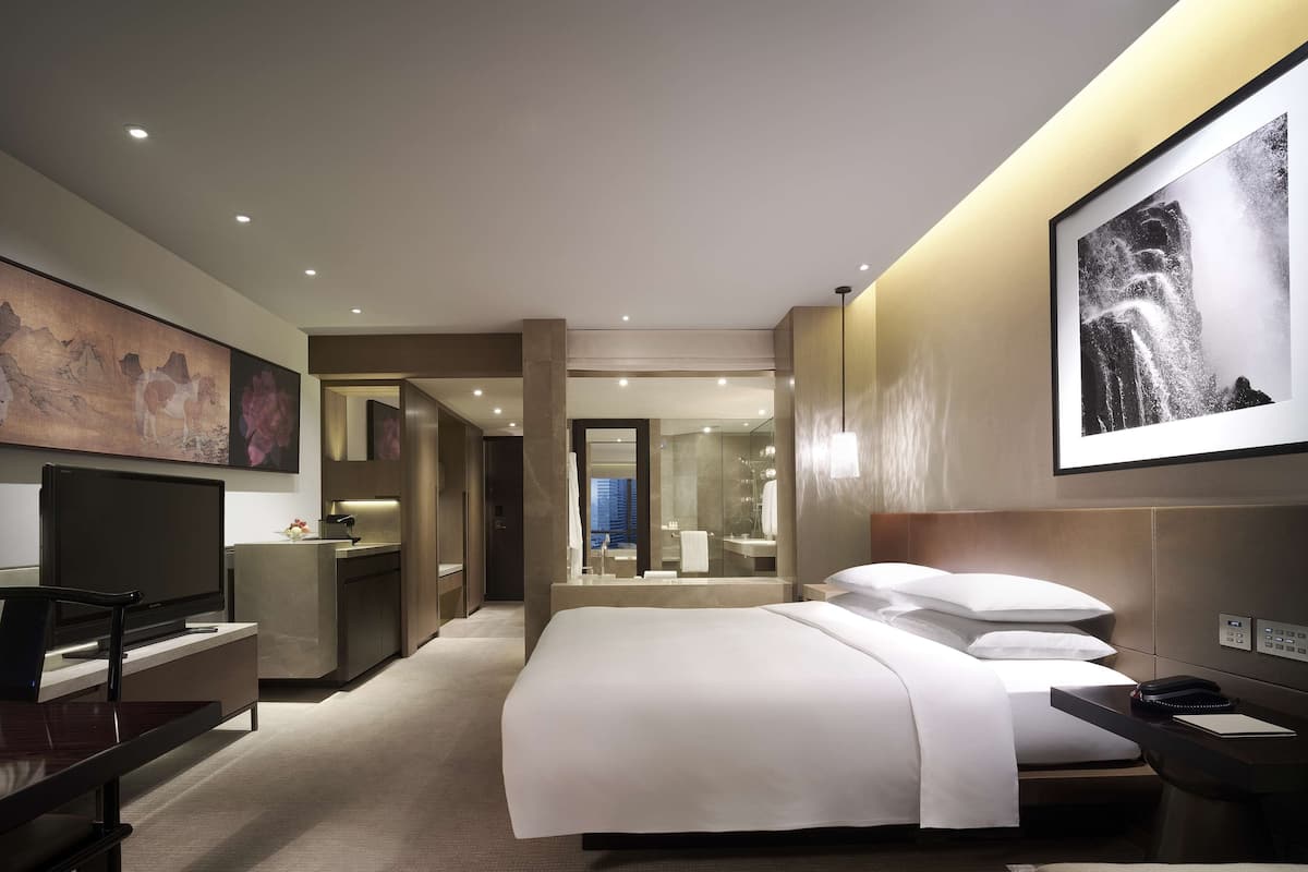 A modern hotel room with a large bed, a desk, a TV, and a bathroom visible through an open door.