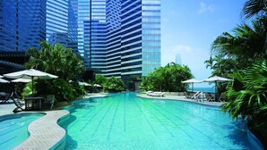 Une piscine entourée de chaises longues et de parasols, bordée de palmiers et de bâtiments modernes de grande hauteur.