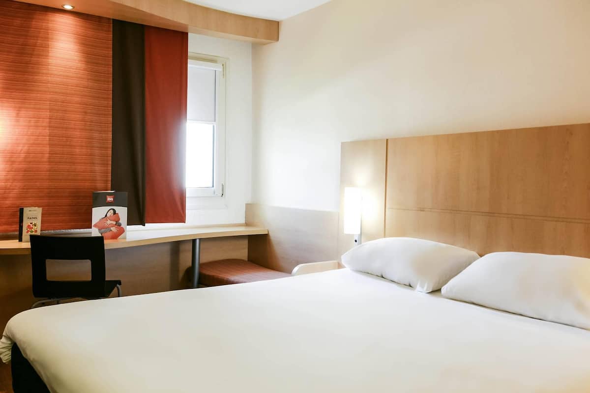 Ein Hotelzimmer mit einem Bett, einem Schreibtisch, einem Sessel, einem Fenster mit Vorhängen und einem Nachttisch mit Lampe.