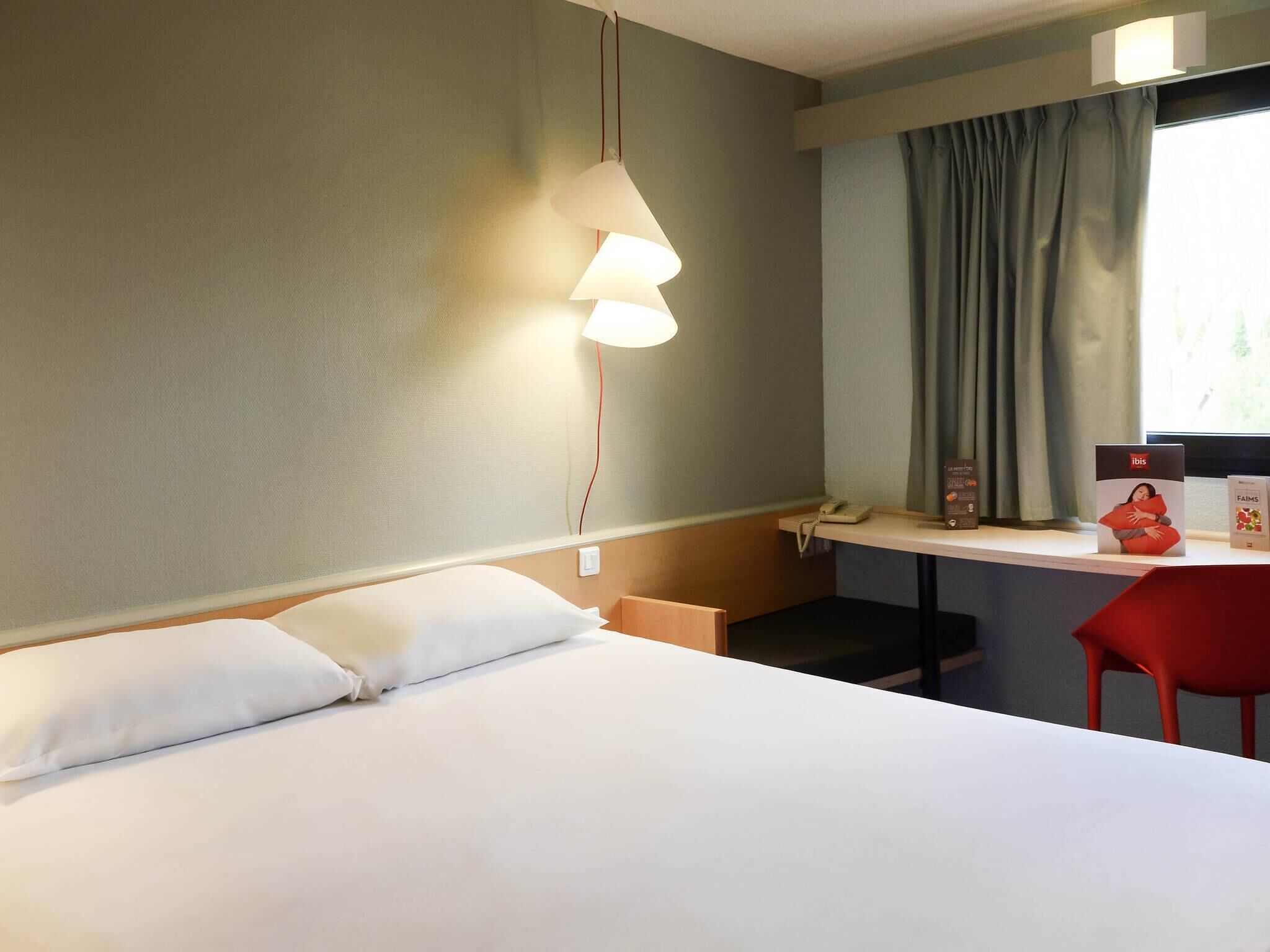 A hotel room with a bed, a desk with a chair, a lamp, and a window with curtains.