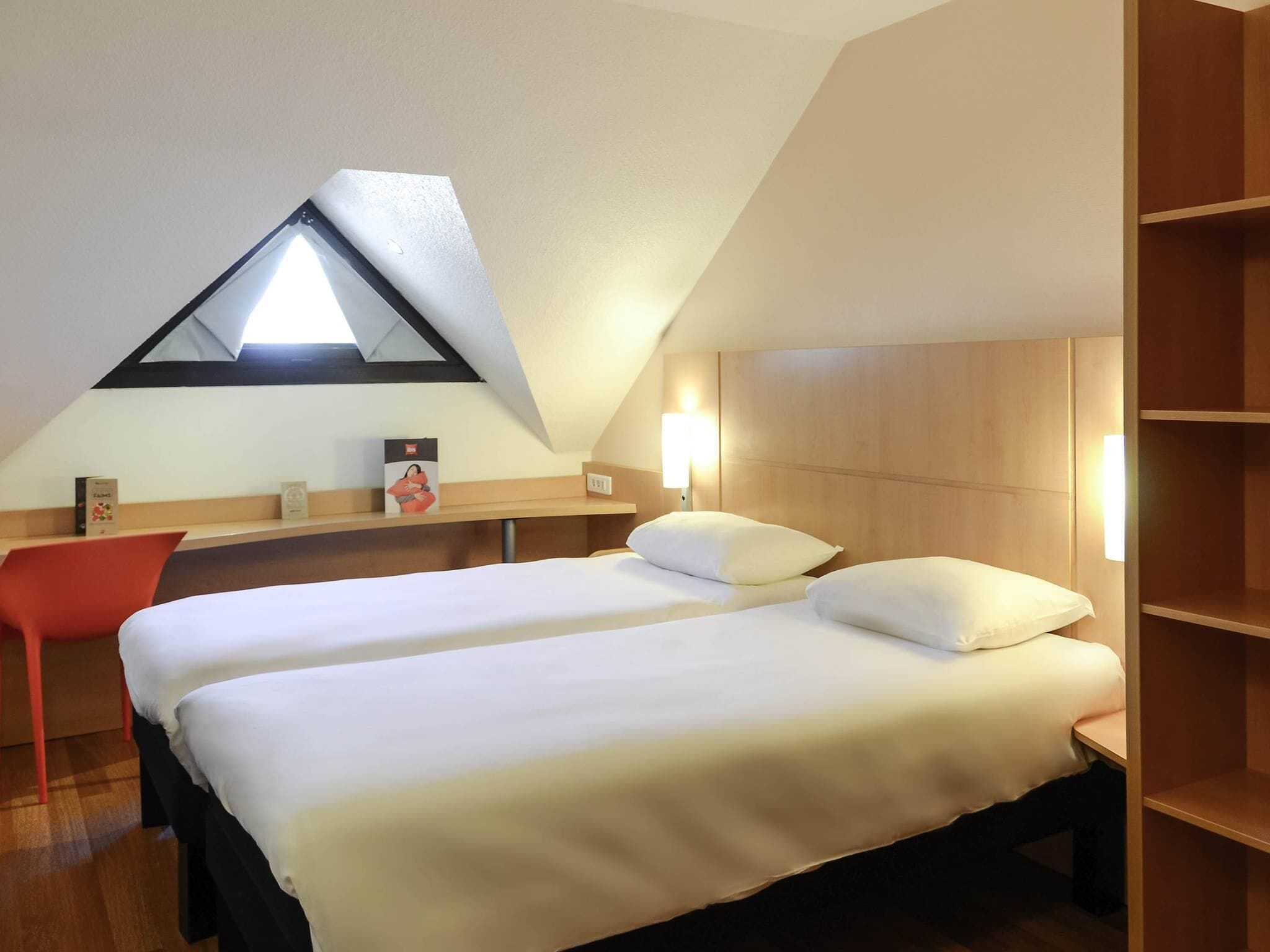 A room with two beds, a red chair, a wooden desk, and a sloped ceiling with a skylight.