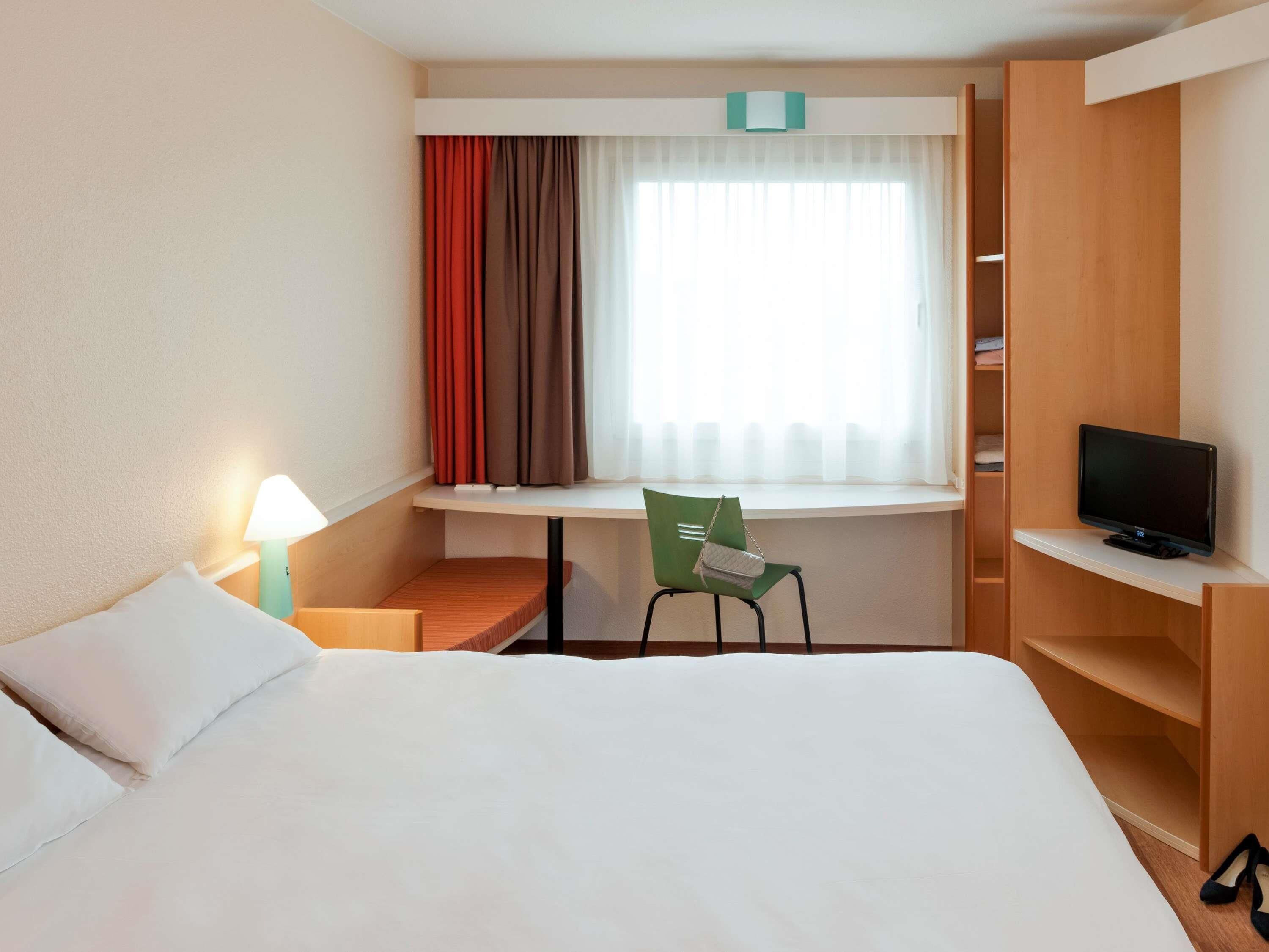 Una habitación de hotel con cama, escritorio con silla, televisión y ventana con cortinas.