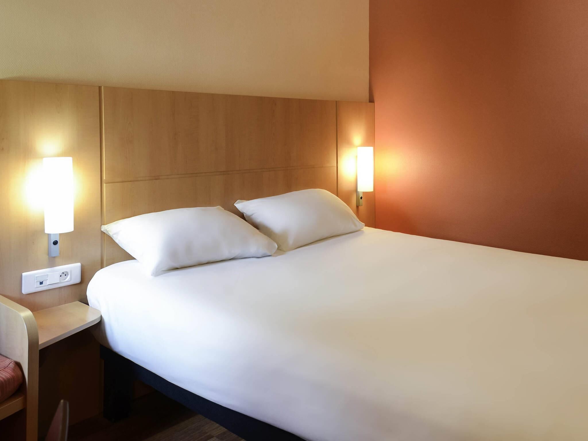 A hotel room with a bed, two bedside lamps, a nightstand, and a power outlet.