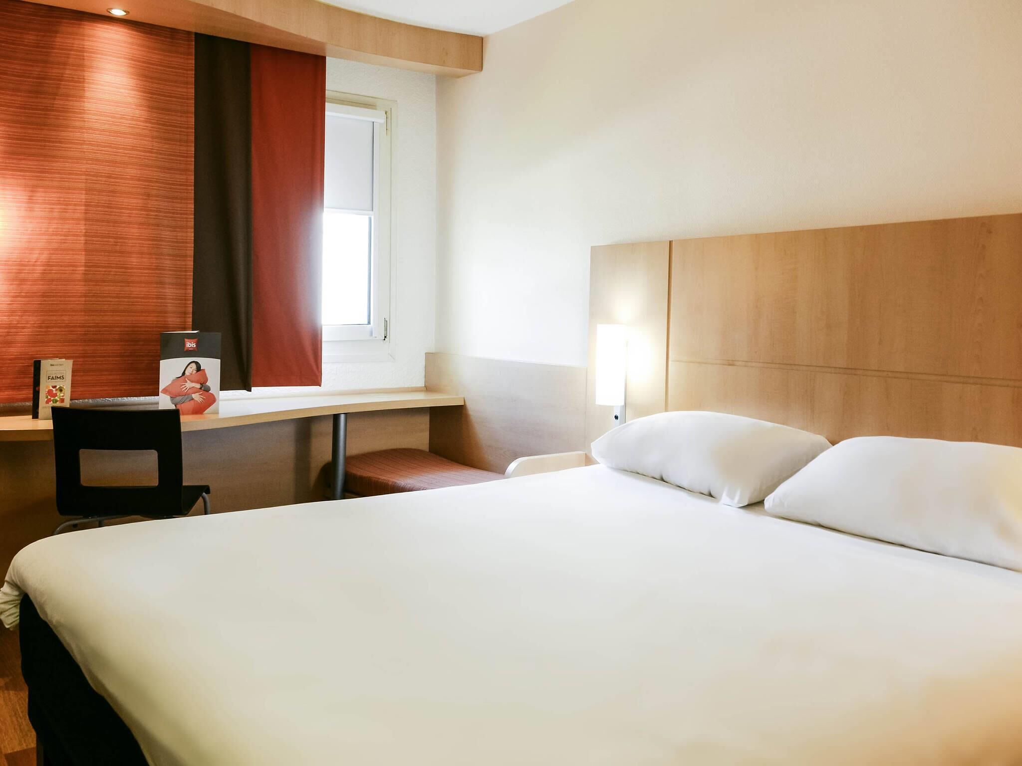 Una habitación de hotel con cama, escritorio, silla, ventana con cortinas y mesita de noche con lámpara.