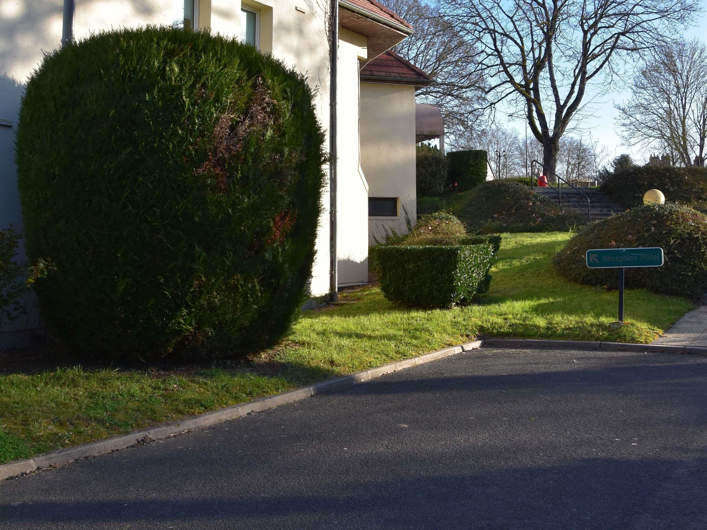 A reception area with a sign for the Récupération Hotel, a building with a white facade, and neatly trimmed hedges.