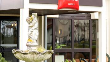 A stone fountain with moss and a statue, in front of a building with a 'ibis Hotels' sign.