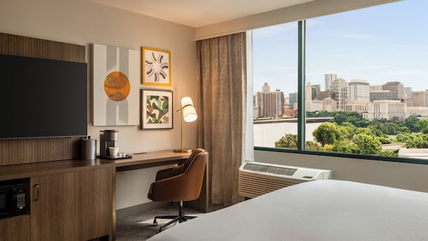 A hotel room with a large bed, a desk with a chair, a flat-screen TV, a coffee maker, and a city view through the window.