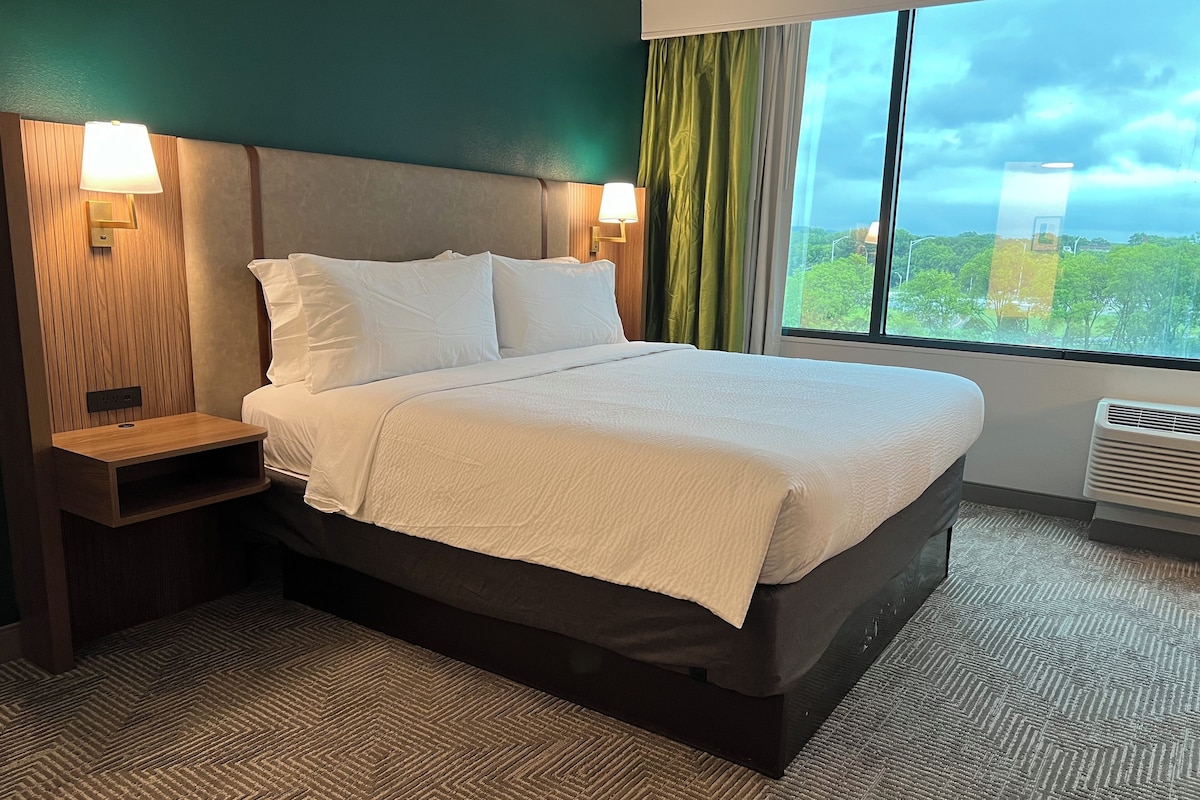 A hotel room with a bed, bedside lamps, a nightstand, a window with curtains, and a view of greenery outside.