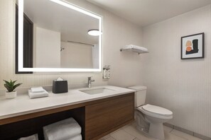 Uma casa de banho moderna com um espelho grande, lavatório branco e sanita. Há um porta-toalhas acima da sanita e um quadro emoldurado na parede.