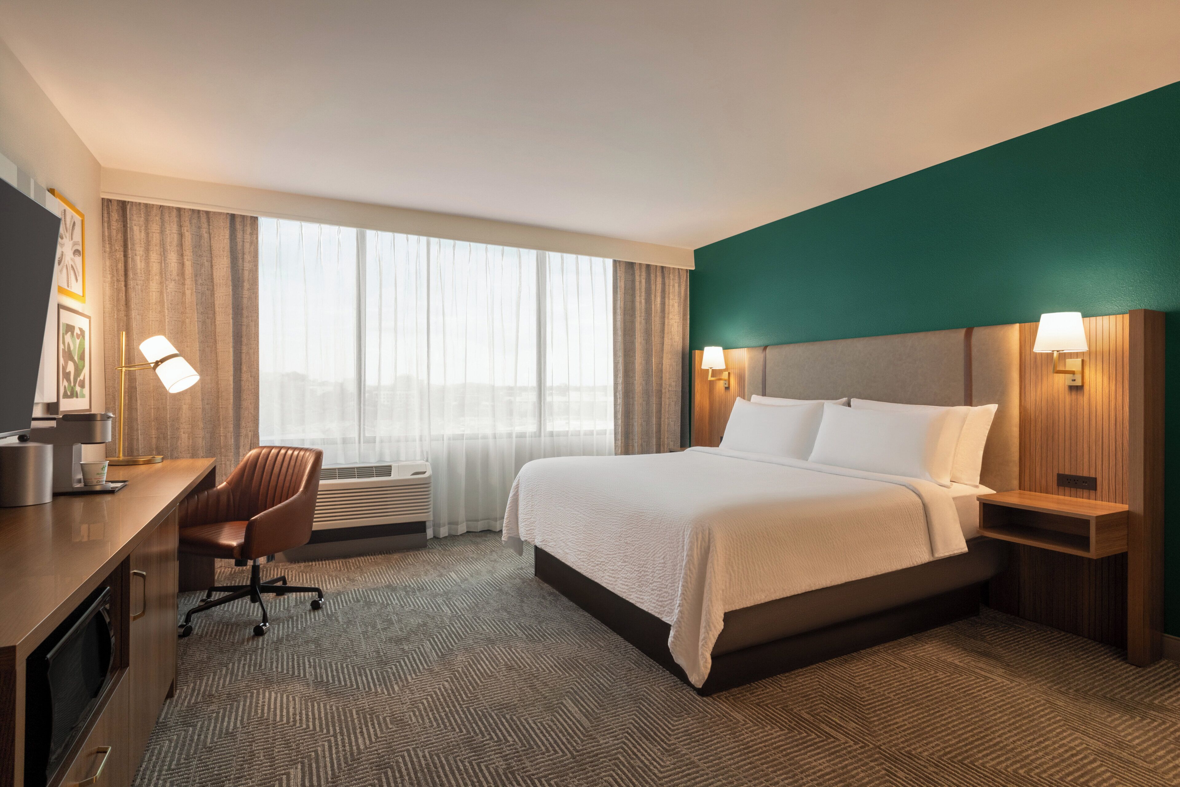 A hotel room with a large bed, a desk with a chair, a TV, and a window with curtains.