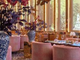 Zona pranzo con sedie rivestite in rosa, un tavolo apparecchiato con bicchieri e piatti, e un vaso azzurro di grandi dimensioni con fiori.