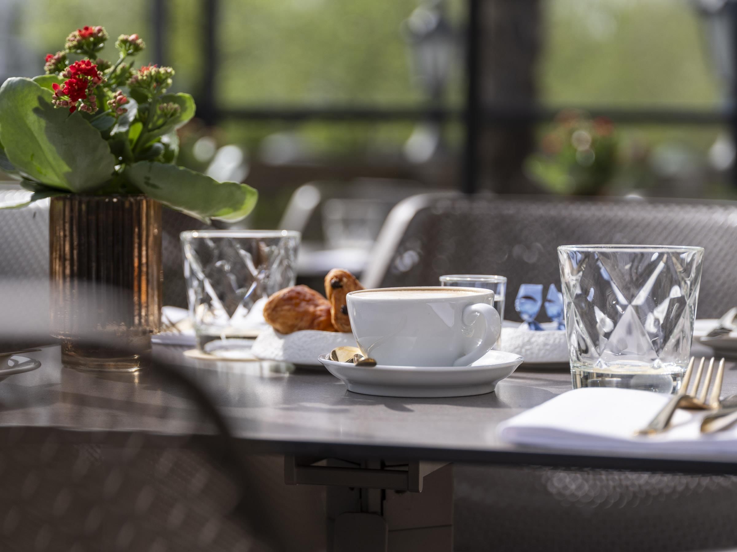 A table set with a cup of coffee, a pastry, and a glass of water, with a vase of flowers in the background.