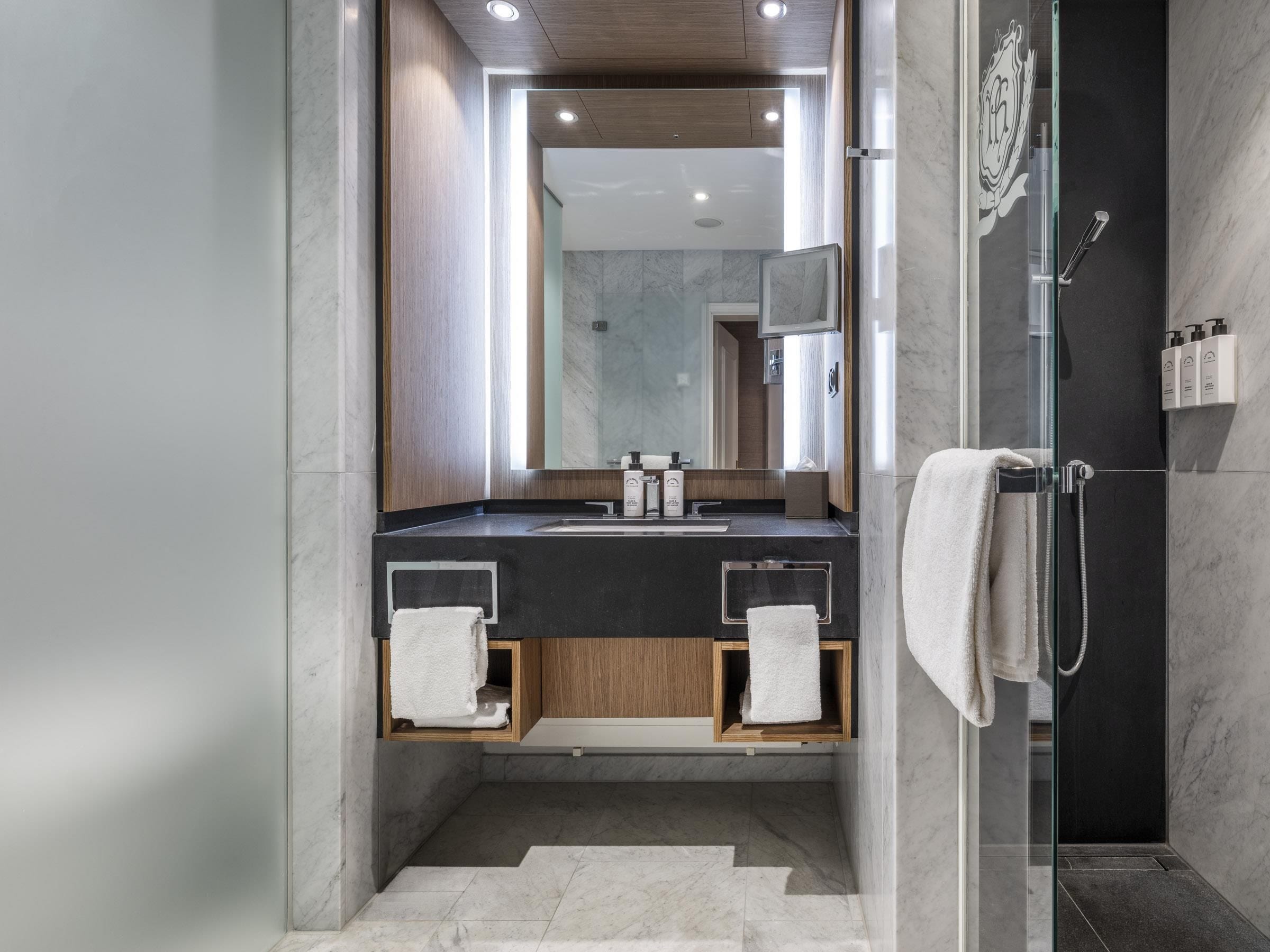 A modern bathroom with a glass-enclosed shower, a marble-tiled floor, and a wooden vanity with a mirror.
