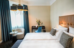 Una camera d'albergo con un letto grande, una scrivania con una sedia, un tavolino con un vaso e una lampada.